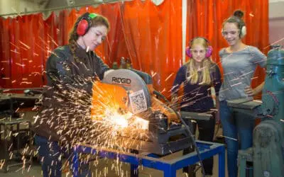 A female engineer cuts metal while two teenage students look on