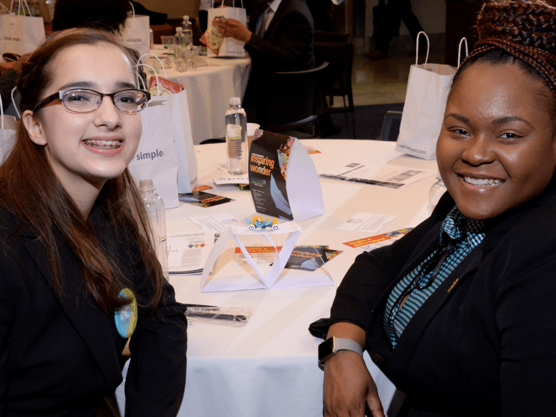 A girl and her mentor sit at a table. They've built a paper structure that can hold up a small toy.