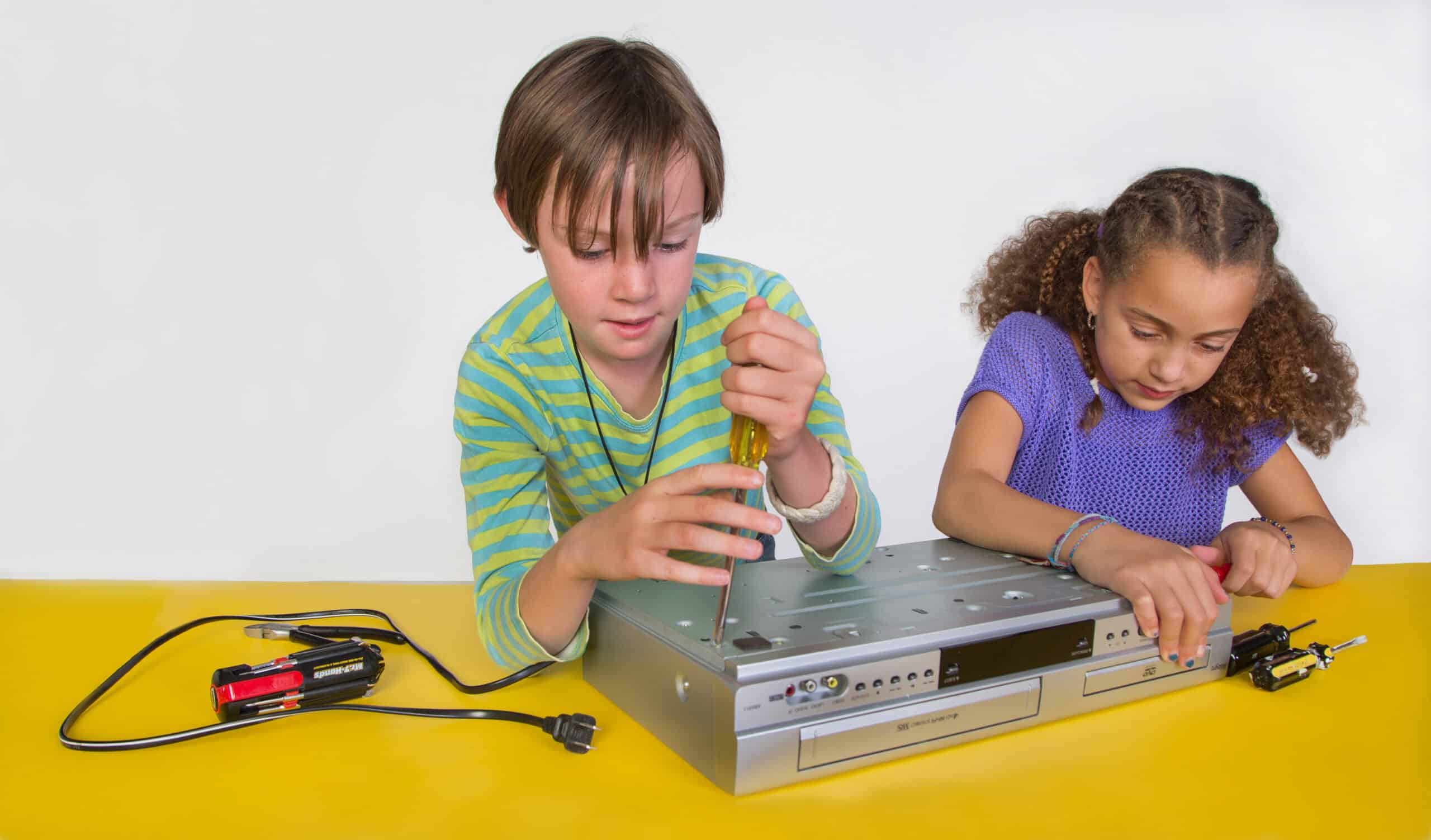 Two middle school girls (one holding a screwdriver) take apart a dvd/vcr machine
