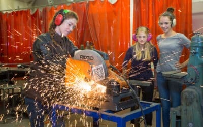 A female engineer cuts metal while two teenage students look on