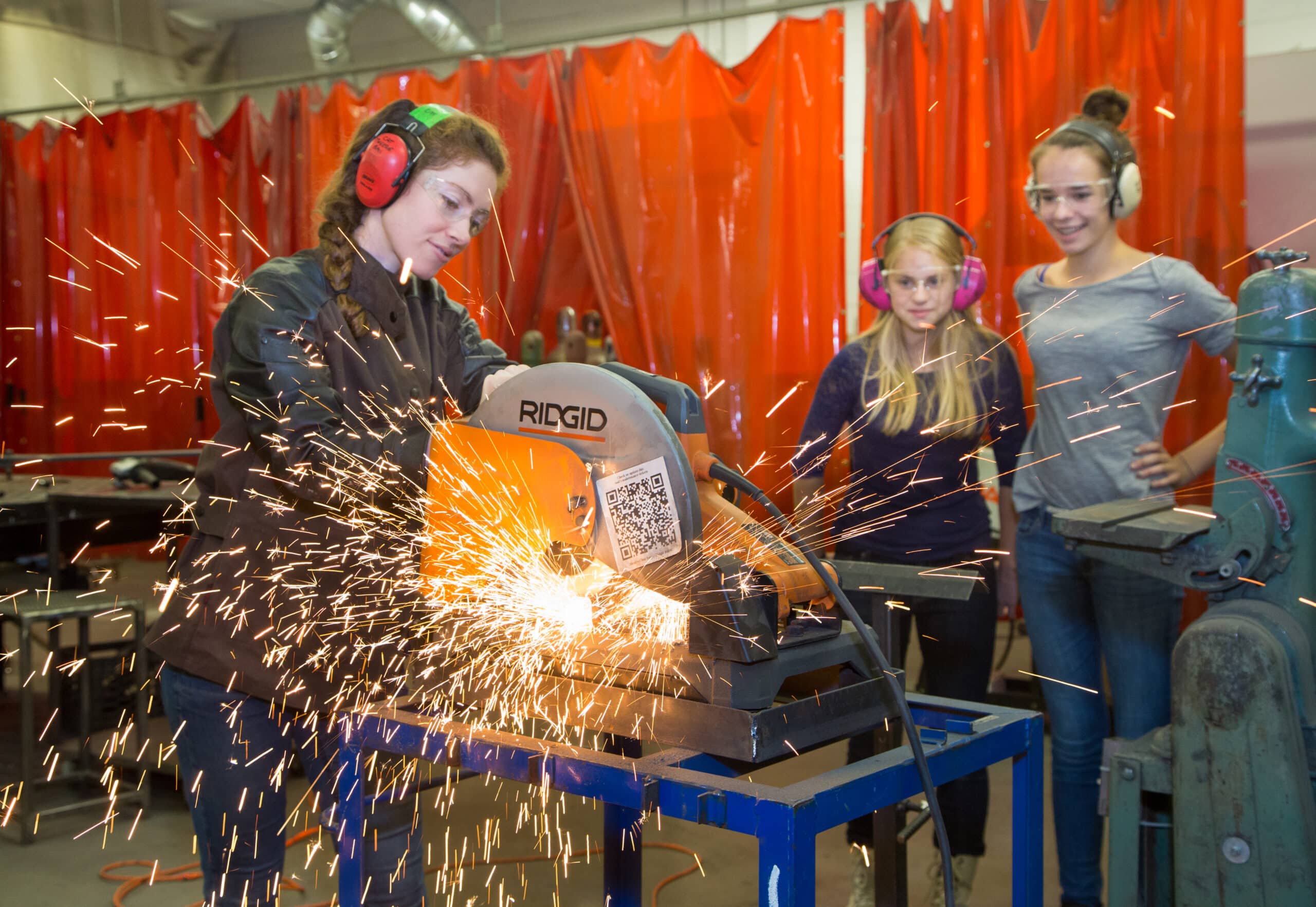 A female engineer cuts metal while two teenage students look on