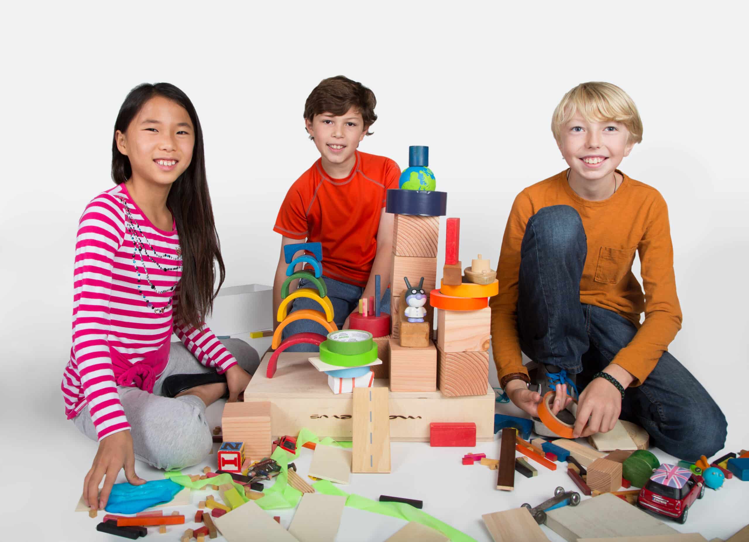 three children look at the camera. They have a pile of recycled products that they can build with.